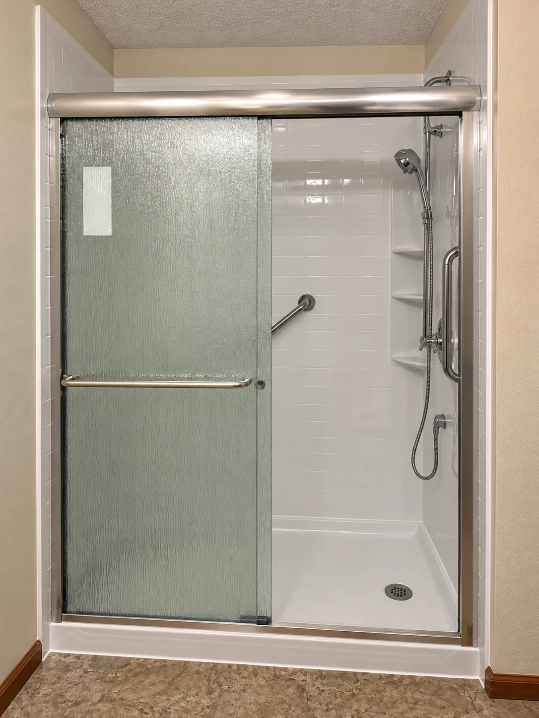 Walk-in shower with white tile-look walls, an angled grab bar and a vertical grab bar, corner shelves, a handheld showerhead on a slide bar, a low-threshold base and a textured sliding glass door.